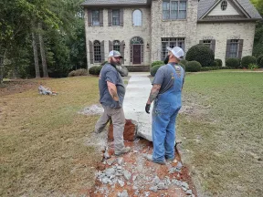 Two workers standing on a broken concrete walkway in front of a brick house preparing for repairs.
