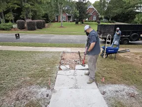 Two workers breaking up a concrete sidewalk in a residential area with tools and a wheelbarrow nearby.