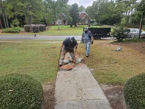 Two workers breaking and removing a concrete sidewalk in a residential front yard with tools and trucks nearby