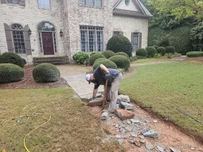 Worker removing broken concrete from front yard walkway repair in residential home with green lawn and bushes.