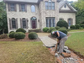 Worker breaking concrete sidewalk outside a large brick house with landscaping and shrubs.