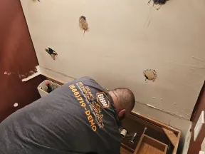 Worker from demolition services repairing bathroom wall with exposed plumbing and damaged drywall.