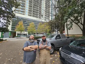 Two bearded men with tattoos stand with arms crossed in an urban outdoor setting near parked vehicles and high-rise buildings.