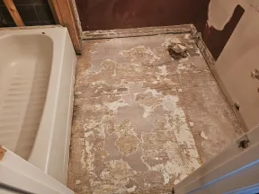 Bathroom floor stripped down to bare plywood next to a white bathtub, showing worn and damaged surface ready for renovation
