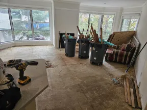 Living room under renovation with removed flooring, trash bins filled with wood debris, and construction tools.