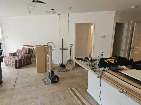 Room under renovation with stripped floors, tools, dolly, striped armchair, and construction materials scattered.