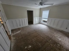 Empty room with bare plywood floor, white wainscoting, beige walls, ceiling fan, and single window with blinds.