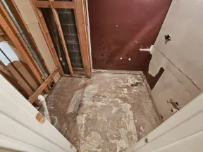 Room under renovation with exposed wooden studs, bare floor, and partially removed wall coverings, plumbing visible.