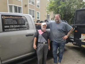 Two men standing beside a demolition service truck in a residential area with company contact info visible.