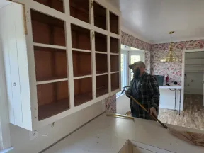 Man with beard and cap working on removing a countertop in a kitchen with floral wallpaper and empty cabinets.