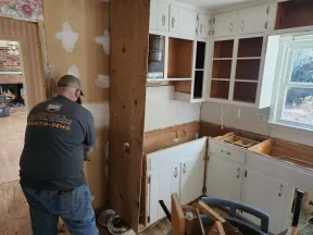 Worker demolishing interior wall in kitchen renovation with white cabinets and open shelving under daylight.