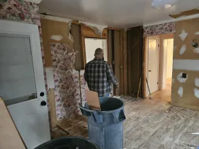 Man renovating a room with torn wallpaper and exposed wall studs during home remodeling project.