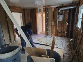 Worker demolishing interior walls and flooring during home renovation with debris and exposed framing visible