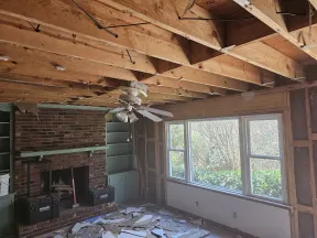 Living room under renovation with exposed ceiling beams, brick fireplace, large window, and debris on floor.