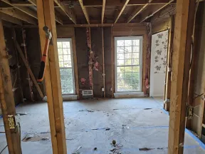 Room under renovation with exposed wooden framing, insulation, two large windows, and concrete floor.