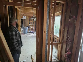 Man in plaid shirt overseeing interior demolition with exposed wooden framing and debris inside house.