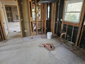 Interior of a house under renovation showing exposed wooden framing, wiring, and plumbing with a window and bucket.