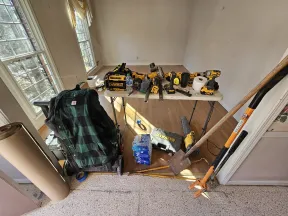 Construction tools and gear including DeWalt drills, shovels, measuring tape, and jacket in a sunlit room with hardwood floor.