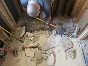 Worker removing old tile flooring and debris in a room under renovation with exposed wooden studs.