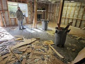 Man stands in a large room under renovation with exposed walls, debris, and trash bins filled with wood scraps.