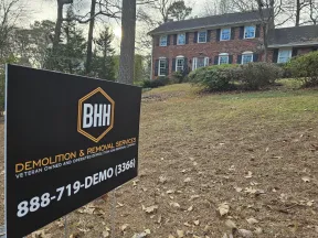 BHH Demolition & Removal Services sign on yard with brick colonial house in background during daytime.