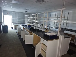 Empty laboratory workspace with open cabinets, black countertops, and shelving under bright fluorescent lighting.