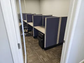 View into an office with empty cubicles and swivel chairs under neutral lighting and beige walls.