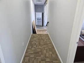 Narrow hallway with patterned carpet tiles and white walls under renovation with construction materials nearby.