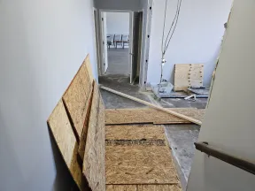 Interior hallway under renovation with plywood sheets, exposed concrete floor, and open door to a room with chairs.