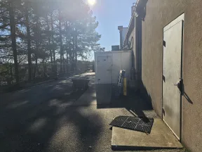 Sunlit industrial alley with metal door, utility equipment, and trees casting shadows on the pavement.