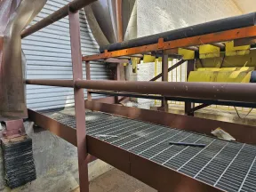 Industrial metal platform with pipes and yellow machinery parts inside a factory setting under bright lighting.