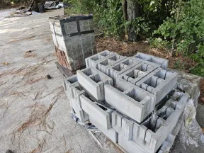 Stacked concrete blocks and bricks on pallets outdoors near wooded area and paved surface.