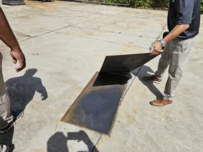 Two men lifting a metal hatch on a concrete surface revealing a dark rectangular underground opening.