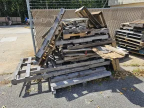 Stack of old wooden pallets and crates discarded outdoors near a chain-link fence under sunlight.