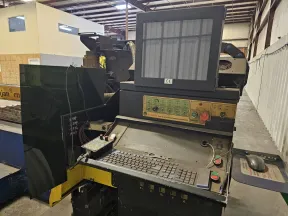 Industrial CNC machine control panel with monitor, keyboard, and emergency stop buttons in factory setting.