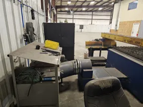 Industrial workspace featuring heavy machinery, ventilation ducts, a worn chair, and metal storage cabinets.
