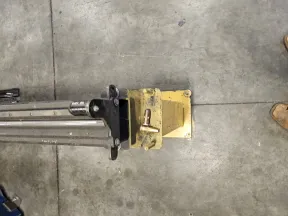 Metal equipment attached to a yellow base on a concrete floor with two bullet casings on top, boots visible at corner.