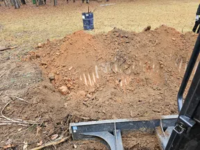 Construction site with excavator digging a large hole in dirt near trees and a trash can in the background.