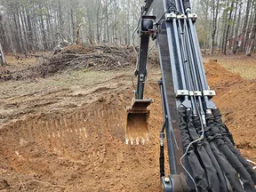 Excavator digging a large hole in a wooded area with dirt piles and branches nearby.