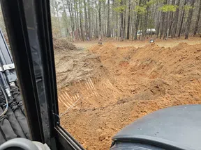 View from construction vehicle cabin showing freshly dug large excavation site surrounded by trees.