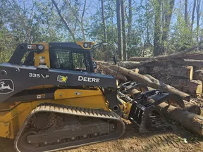 John Deere 335P tracked skid steer loader moving large logs in a forest clearing under blue sky.