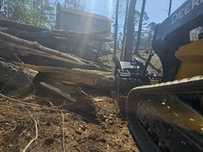 John Deere construction equipment clearing large cut tree logs near a residential house on a sunny day.
