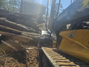 John Deere construction vehicle with a bucket loader moving large tree logs near a house on a sunny day.