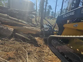 John Deere skid steer loader moving large tree logs in a wooded area with a house in the background.