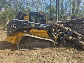John Deere 335 skid steer loader clearing logs in a forested area with tracked wheels and grapple attachment.