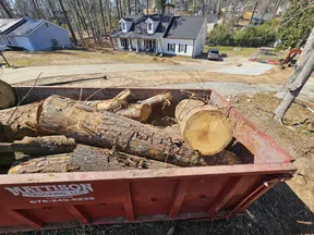 Large red waste container filled with cut tree logs near residential houses on a sunny day