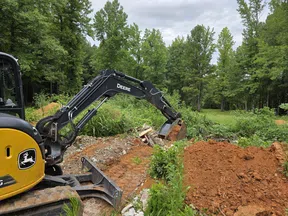 John Deere excavator digging dirt at a construction site surrounded by trees and greenery