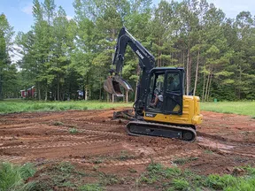 Yellow and black John Deere excavator digging on red dirt clearing surrounded by green trees under blue sky.