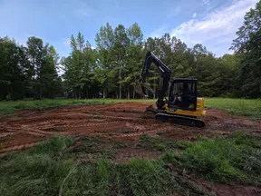 Yellow excavator digging soil in a forest clearing with green trees and blue sky in the background.
