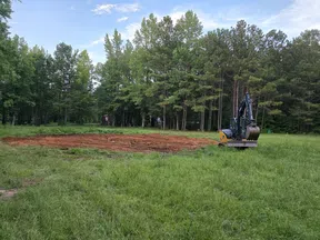 Mini excavator digging a large dirt area in a grassy field surrounded by trees under a blue sky
